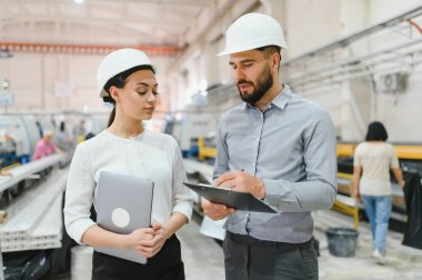 Kask takan iki mühendis, ellerinde bir dizüstü bilgisayar ve bir pano olan plastik pencere fabrikasında çalışmayı tartışıyorlar.