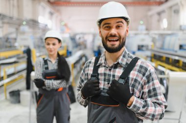Modern bir endüstriyel fabrikada makinelerin önünde gülümseyen işçi tulumları ve kasklar takan iki fabrika işçisi