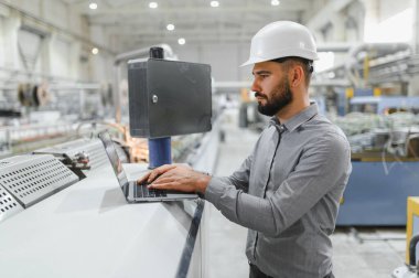 Endüstriyel mühendis sabit şapka takıyor ve laptop kullanıyor. Modern bir imalat tesisinde operasyonları denetliyor.