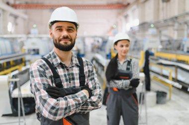 Tulumlu ve kasklı iki fabrika işçisi büyük bir sanayi bölgesinde kollarını kavuşturarak ayakta duruyorlar.