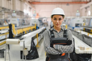 Güvenlik kaskı ve üniforması giyen kendine güvenen kadın fabrika işçisi üretim hattı makinelerinin önünde silah çekerek endüstriyel ortamdaki uzmanlık ve profesyonelliği gösteriyor.