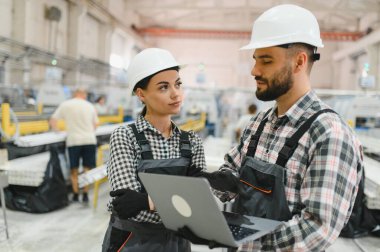Sert şapka ve tulum giyen iki fabrika işçisi, fabrika zeminindeki üretimi tartışmak için dizüstü bilgisayar kullanıyorlar.