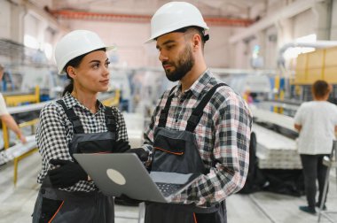 Güvenlik kaskı ve tulum giyen iki fabrika işçisi, yoğun bir fabrika ortamında üretim hattı optimizasyonunu tartışan bir dizüstü bilgisayar kullanıyorlar.