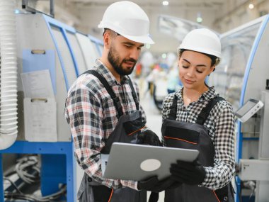 Tulum ve şapka giyen iki fabrika işçisi bir dizüstü bilgisayar kullanıyor, modern bir endüstriyel tesiste üretim süreçlerini tartışıyorlar.