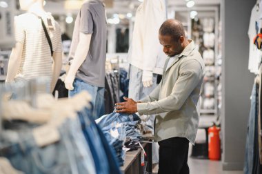 Bir perakende mağazasından kot pantolon seçen şık bir adam, çeşitli giysiler sergileyen mankenlerle çevrili