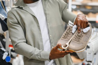 Bir perakende mağazasında gezinirken şık bir adam elinde bej renkli bir çift spor ayakkabı tutuyor, şık bir alışveriş deneyiminin tadını çıkarıyor.