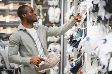 Ayakkabı mağazasında alışveriş yaparken çeşitli spor ayakkabılarını karşılaştıran şık bir adam, rahatlık, tasarım ve güncel moda eğilimlerini göz önünde bulundurarak
