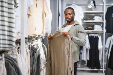 Bir perakende mağazasında giysi seçeneklerini gözden geçirirken bej gömlek tutan şık bir adam, keyifli bir alışveriş deneyimi yaşıyor.