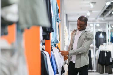 Afro-Amerikalı bir adam alışveriş merkezindeki kıyafetleri karıştırıyor, şık kıyafetler seçmeye odaklanıyor. Sahne bir moda ve seçim anını yakalar.