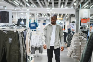 Genç, şık, Afro-Amerikalı bir adam giyim mağazasından geçiyor ve alışveriş merkezinden kıyafet seçiyor.