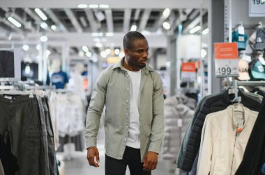 Delikanlı bir moda mağazasında geziniyor, yeni şık kıyafetler ararken askılardaki kıyafetleri dikkatle inceliyor.