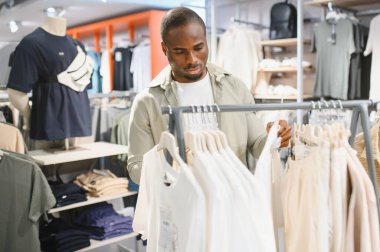 Giysi rafından gömlek seçen, modern alışveriş merkezlerinde alışveriş yapan, odaklanmış Afro-Amerikan bir adam.
