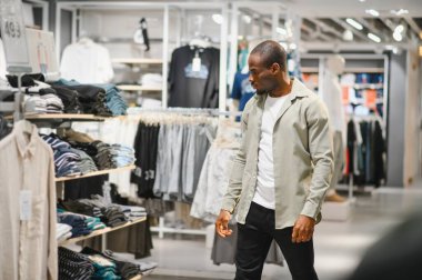 Afro-Amerikalı bir adam alışveriş merkezindeki elbise raflarını karıştırıyor, şık kıyafetler seçiyor. Moda ve kişisel stile odaklan.