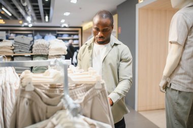 Alışveriş merkezindeki bir giyim mağazasında Afro-Amerikalı bir adam raftan şık kıyafetler seçiyor ve moda seçimlerine odaklanıyor.