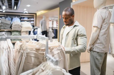 Afro-Amerikalı şık bir adam perakende mağazasında kıyafet seçiyor, sergilenen çeşitli kıyafetleri dikkatlice inceliyor.
