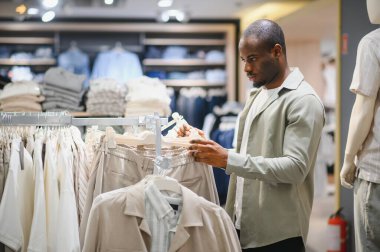 Tarz sahibi Afro-Amerikan erkeği modern moda mağazasında giysileri dikkatle seçiyor, modern alışveriş merkezlerinde alışveriş deneyiminin tadını çıkarıyor.