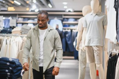 Modern bir alışveriş merkezinde mankenler ve çeşitli giysilerle çevrili bir perakende mağazasında kıyafet seçen şık bir Afro-Amerikan erkeği.