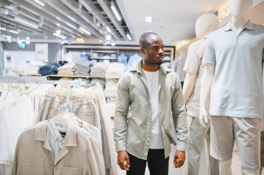 Modern, hareketli bir alışveriş merkezinde alışveriş yaparken kıyafetleri karıştıran ve mankenleri inceleyen şık bir adam.