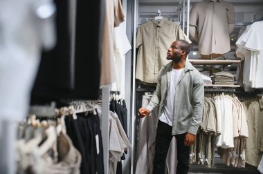 Afrika kökenli Amerikalı bir adam alışveriş merkezinde kıyafet seçeneklerini gözden geçiriyor. Modern bir gardırop güncellemesi için moda seçimlerine odaklanıyor.