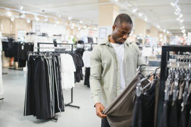 Genç Afro-Amerikalı bir adam bir giyim mağazasında yeni pantolon almak için alışverişe çıkıyor, bir pantolon giyiyor ve elbise askılarına bakıyor.