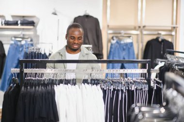 Gülümseyen Afrikalı Amerikalı bir adam perakende mağazasında kıyafet seçiyor, alışveriş deneyiminden zevk alıyor.