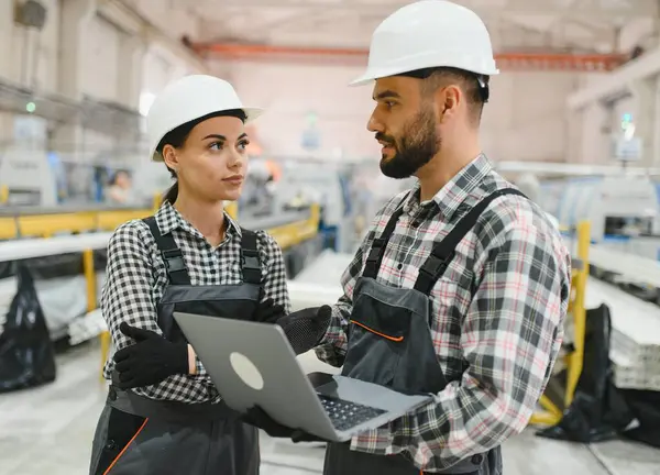 İki fabrika işçisi, bir erkek ve bir kadın, büyük bir endüstriyel binada dizüstü bilgisayar tutarken üretimi tartışıyorlar.