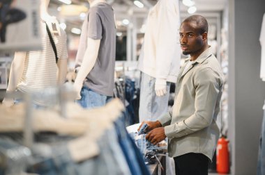 Müşteri moda mağazasına göz gezdiriyor, yeni kot pantolon seçiyor ve son moda modellerle çevrili.