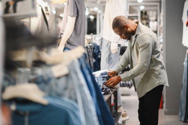 Bir giyim mağazasında kot pantolon alışverişi yapan Afro-Amerikalı bir adam farklı tarz ve renkleri inceliyor.