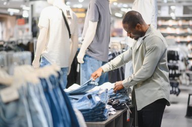 Müşteri, mankenler ve çeşitli kıyafetlerle çevrili bir mağazadan kot pantolon seçiyor.