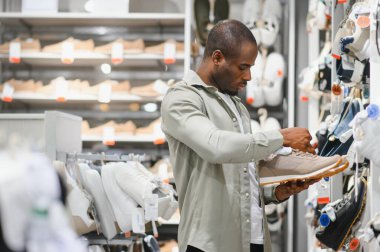 Ayakkabı mağazasında alışveriş yaparken bir çift bej ayakkabıyı inceleyen bir adam, çeşitli ayakkabı seçenekleriyle çevrili.