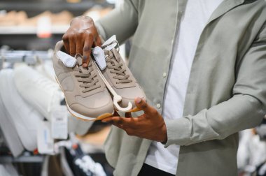 Bir perakende mağazasında dolaşırken şık bir adam elinde bej bir çift spor ayakkabı tutuyor, düşünceli seçimler yapıyor.