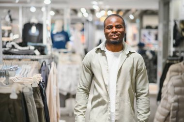 Afro-Amerikalı bir adam alışveriş merkezinin giyim mağazasında moda seçeneklerini araştırıyor. Modern bir perakende satış tarzı ve seçimi sergiliyor.