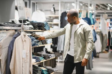Afrikalı Amerikalı bir adam modern bir alışveriş merkezinde moda ve moda tercihlerine odaklanarak kıyafetleri karıştırıyor ve seçiyor.