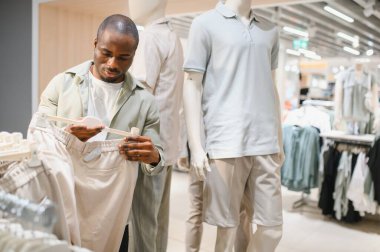 Afro-Amerikalı bir adam alışveriş merkezinde moda seçeneklerini araştırıyor. Mankenlerle çevrili bir raftan şık kıyafetler seçiyor.