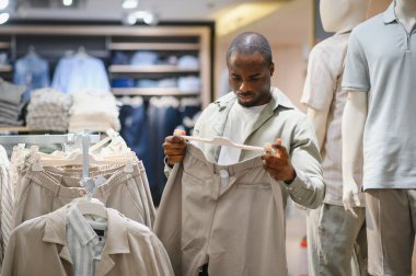 Modern bir giyim mağazasında alışveriş yaparken askıdaki pantolonu dikkatle inceleyen şık bir Afro-Amerikan erkeği.