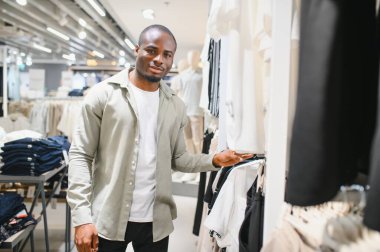 Modern bir alışveriş merkezinde giyinme raflarını karıştıran Afrikalı Amerikalı bir adam moda kıyafetlerini güncellemek için moda ve moda tercihlerine odaklanıyor.