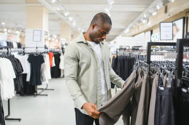 Genç Afro-Amerikalı bir adam bir giyim mağazasında yeni pantolon almak için alışverişe çıkıyor. Satın almadan önce kumaşı ve stili dikkatlice inceliyor.