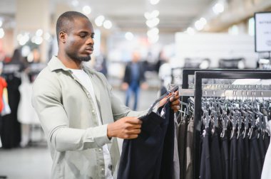 Genç adam alışveriş deneyiminin tadını çıkarırken bir perakende mağazasında şık giysi ürünleri seçiyor ve son modaya özenle göz atıyor.