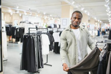 Gösterişli bir moda mağazasında alışveriş yaparken şık bir pantolon tutan müşteri, çeşitli moda kıyafetleri ve aksesuarlarla çevrili.