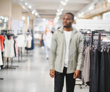 Kendine güvenen Afrikalı Amerikalı adam elbise raflarında yürüyor, modern alışveriş merkezlerinde alışveriş deneyiminin tadını çıkarıyor.