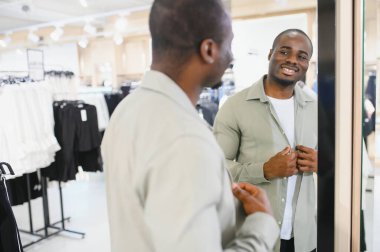 Gülümseyen müşteri aynanın önünde şık kıyafetler deniyor, canlı bir giyim mağazasında alışveriş deneyiminin tadını çıkarıyor.