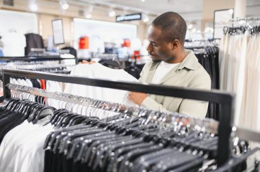 Müşteri beyaz tişörtü inceliyor elbise askısına asılıyor, modern giyim mağazasından yeni kıyafetler alıyor.