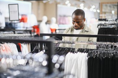 Alışveriş merkezinde alışveriş yapan, raflardan şık kıyafetler seçen, modern moda tercihlerini ve kişisel tarzını sergileyen bir adam.