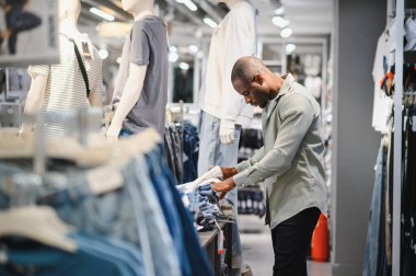 Genç Afro-Amerikalı bir adam giyim mağazasından kot pantolon alıyor, farklı stil ve renkleri inceliyor.