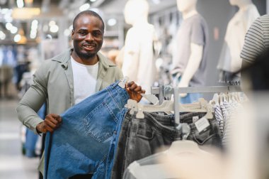 Kot tutan şık bir adam, bir mağazada kıyafet seçiyor ve karşılaştırıyor, alışveriş deneyiminin tadını çıkarıyor.
