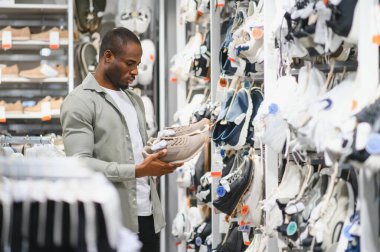 Genç Afro-Amerikalı bir adam seçeneklerle dolu geniş bir ayakkabı mağazasında gezinirken bir çift moda bej spor ayakkabı seçiyor.
