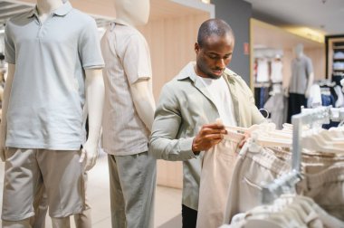 Şık bir Afro-Amerikan erkeği, mankenler ve çeşitli kıyafetlerle çevrili bir giyim mağazasında pantolon seçerken dikkatle seçiyor.