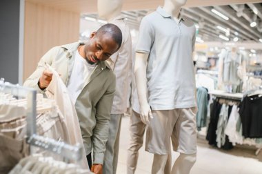 Şık bir Afro-Amerikan erkeği modern moda mağazasında giysileri dikkatlice seçiyor, kumaşları inceliyor ve tarzları karşılaştırıyor.