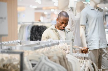 Mankenler ve şık giysi vitrinleriyle çevrili modern bir alışveriş merkezindeki kıyafetleri özenle seçen Afrikalı Amerikalı adam moda odaklı...