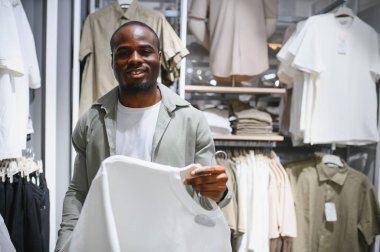Gülümseyen Afrikalı Amerikalı müşteri elbise mağazasında askıda beyaz t-shirt tutuyor, moda kıyafet alışverişinin keyfini çıkarıyor.
